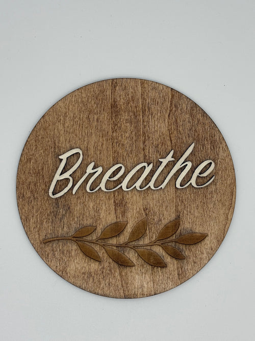 Wooden circular sign with 'Breathe' and leaf design on a light gray background