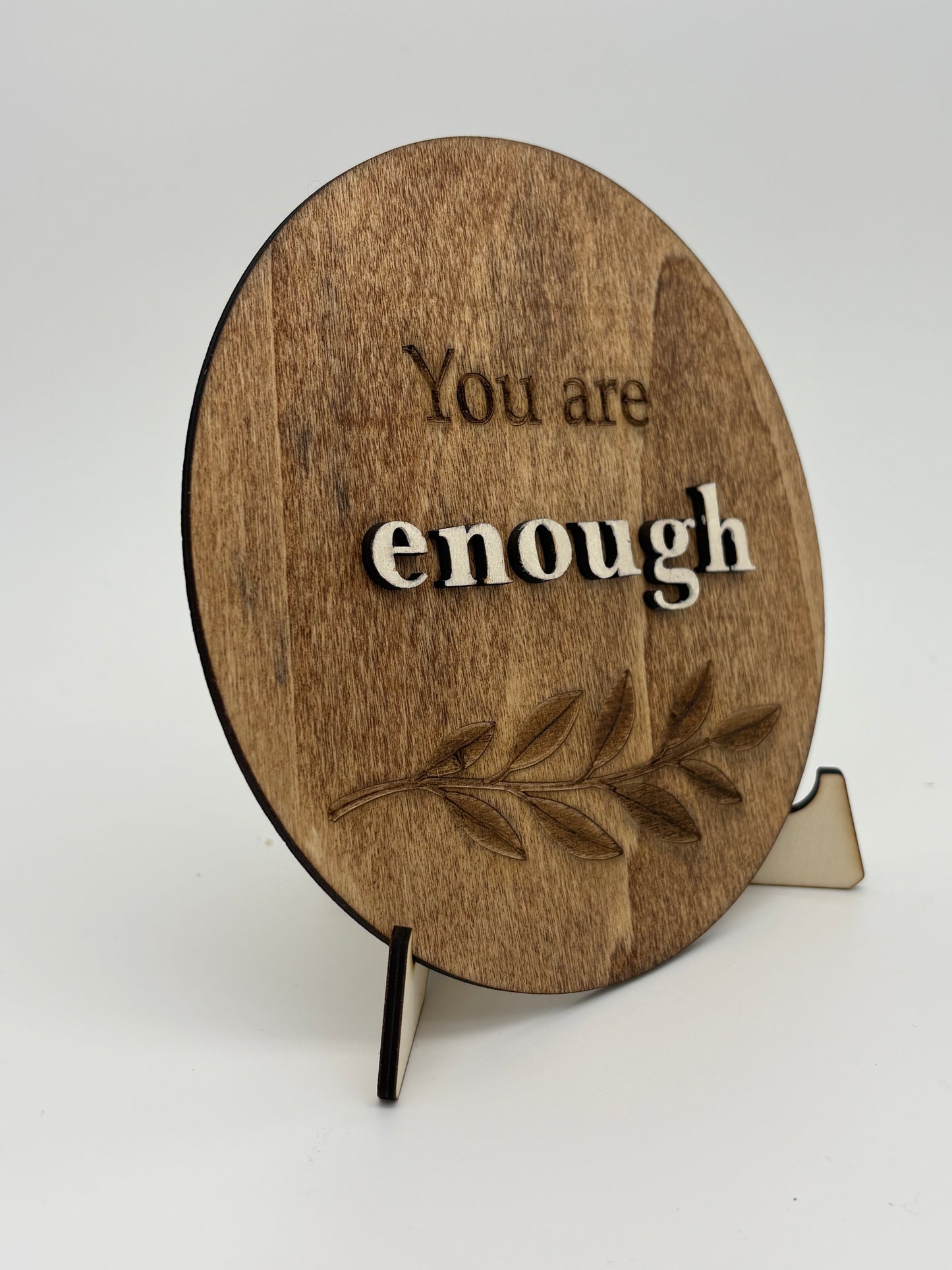 Wooden circular sign with 'You are enough' text and a leaf design on a stand.