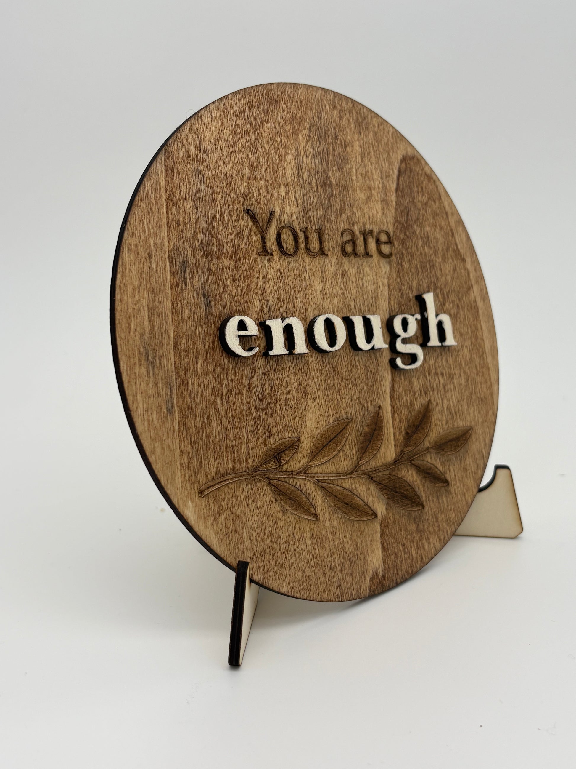 Wooden circular sign with 'You are enough' text and a leaf design on a stand.