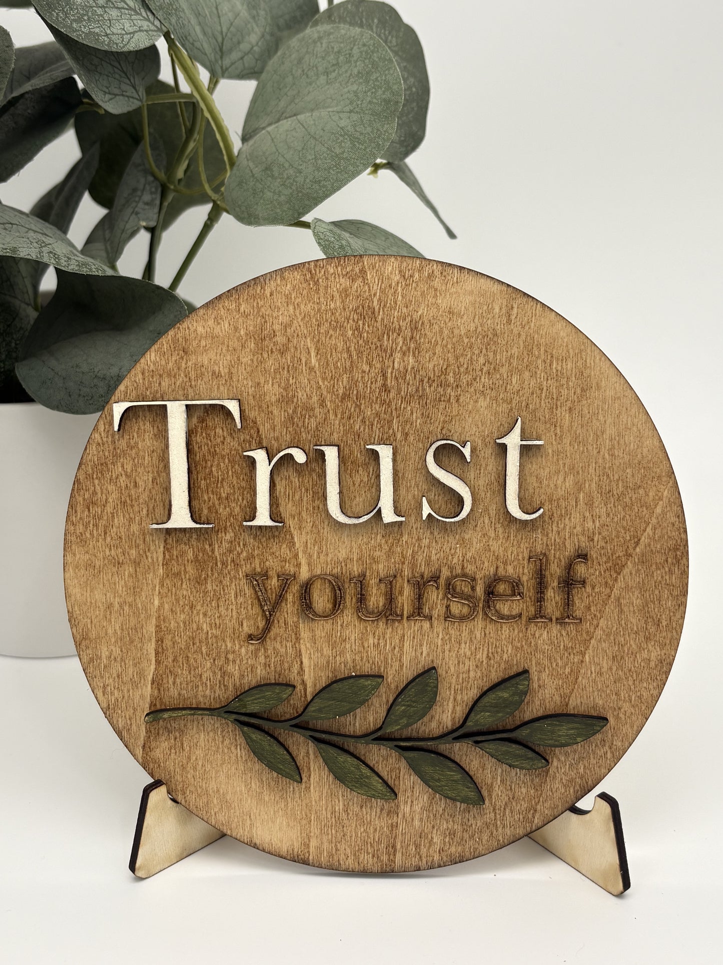 Wooden circular sign with 'Trust yourself' text and leaves on a stand.